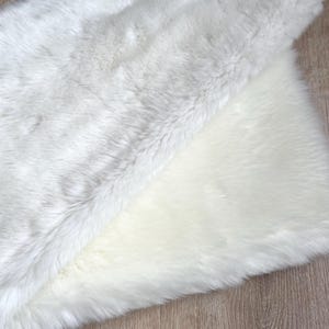 May include: A white faux fur rug with a soft, fluffy texture. The rug is folded in half, showing the underside of the fur.