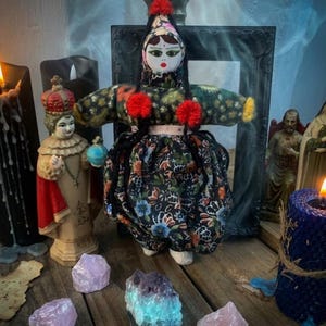 May include: A handcrafted doll with a floral dress and arms, decorated with red pom-poms. The doll is surrounded by crystals, candles, and religious figurines, creating a mystical and spiritual aesthetic.