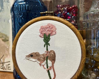May include: Embroidered hoop art featuring a brown mouse climbing a green stem with a pink flower at the top.