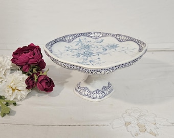 May include: A white ceramic pedestal cake stand featuring a blue floral pattern in the center and a blue lace-like decorative border along the scalloped rim and the base. The stand has a smooth, glossy finish and a wide, shallow circular plate.