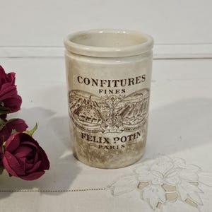 May include: A cylindrical ceramic jar with a cream-colored base and a distressed finish. The front features dark brown text reading CONFITURES FINES and FELIX POTIN PARIS, along with a detailed illustration of a factory building complex.
