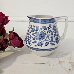 Peut inclure: Une cruche en céramique blanche avec un motif floral bleu. La cruche a une anse et un bec verseur. Le motif floral est un dessin répété de feuilles et de fleurs. La cruche est posée sur une surface blanche.