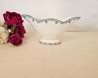 May include: A white ceramic gravy boat with a green and white geometric pattern around the rim and base. The boat has a scalloped edge and a flared base.