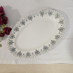 Puede incluir: Plato ovalado blanco con un borde floral decorativo negro y gris. El plato vacío está sobre una superficie blanca. Un ramo de flores rojas oscuras y blancas está en la esquina superior izquierda.