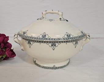 May include: Cream-colored, lidded serving dish with a pedestal base and handles. The dish is adorned with a blue floral and leaf pattern around the rim, lid, and base. The handle on the lid is arched. A decorative piece for serving food.
