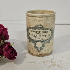 May include: A cylindrical beige ceramic jar with a mottled finish featuring a dark green decorative label that reads CONFITURES FINES FELIX POTIN PARIS. The jar is positioned next to a cluster of deep magenta and white artificial flowers.