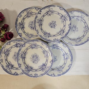 May include: Six white ceramic plates with a blue floral pattern. Each plate has a central floral bouquet and a border of blue flowers and ribbons. The plates are arranged on a white surface, with flowers in the upper left corner.