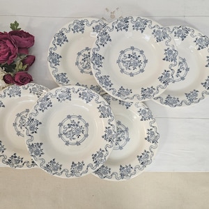 May include: Six white ceramic plates with a blue floral pattern. Each plate features a scalloped edge and a central floral design. The plates are displayed on a white surface, with flowers in the background.