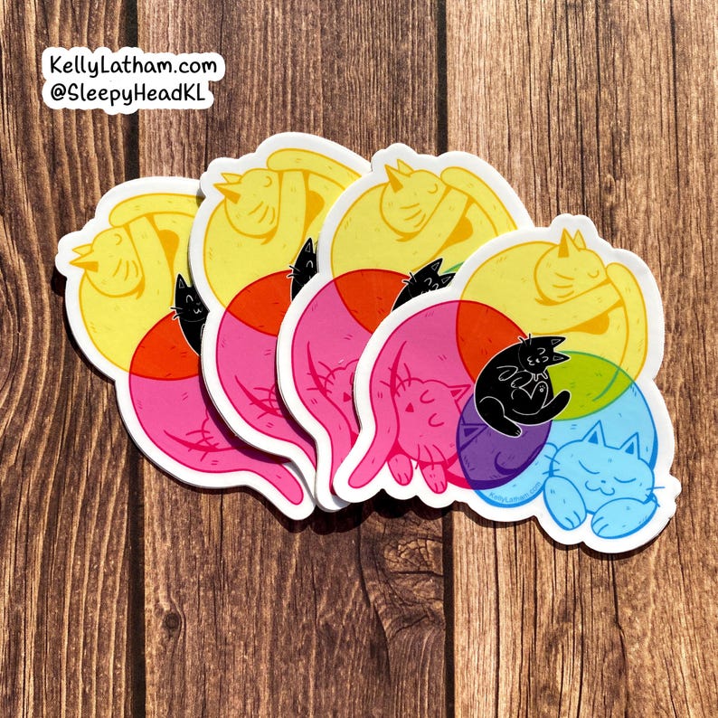 May include: Five colorful stickers featuring a cat sleeping in a circle with a smaller cat inside. The stickers are layered on top of each other, with the bottom sticker being blue, the next being purple, then pink, yellow, and finally yellow again. The stickers are on a wooden surface.