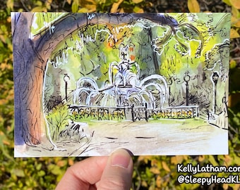 May include: Watercolor and ink illustration of a park scene featuring a fountain with water jets. The artwork includes a tree, lampposts, and a decorative fence. The artist's signature and social media handle are visible in the bottom right corner.