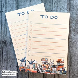 May include: Two cream-colored to-do list notepads with blue and orange lines. The bottom of each notepad features a stack of books with a cat peeking out from the top.