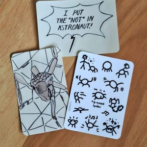 May include: Three rectangular stickers with white backgrounds and black ink illustrations. One features a speech bubble with the text I PUT THE "NOT" IN ASTRONAUT!. Another shows a spider-like creature, and the third displays various stick-figure spider doodles with the text He is a big-assed spider.