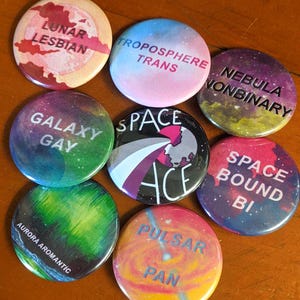 May include: Nine round buttons with space-themed designs and text. The buttons feature various colors, including pink, blue, green, purple, and black. The text on the buttons includes "Lunar Lesbian", "Troposphere Trans", "Nebula Nonbinary", "Galaxy Gay", "Space Ace", "Space Bound Bi", "Aurora Aromantic", "Pulsar Pan".