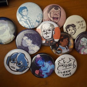 May include: A collection of round, colorful buttons featuring cartoon illustrations of characters. The buttons display various characters in different poses and expressions, with some including text like "Treat Yo Self" and "Sleep Time". The buttons are arranged on a wooden surface.