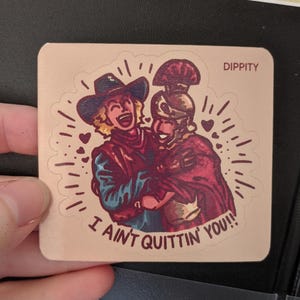 May include: A sticker featuring a cartoon cowboy and a Roman soldier embracing. The cowboy is wearing a brown hat and a blue shirt. The Roman soldier is wearing a red cape and a helmet. The sticker has the text "I ain't quittin' you!" and "Dippity" in the top right corner.