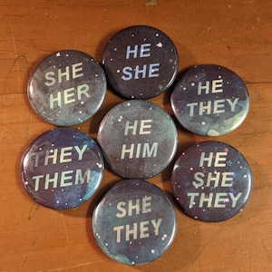 May include: Eight round buttons with a galaxy-like background. Each button has a different combination of the words "He", "She", "They", "Him", and "Them" in white text.