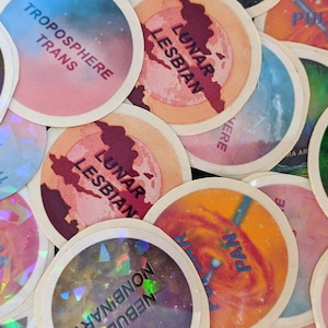 May include: A collection of colorful stickers with various space-themed designs and text, including "Space Ace", "Lunar Lesbian", "Troposphere Trans", "Galaxy Gay", and "Nebula Nonbinary".