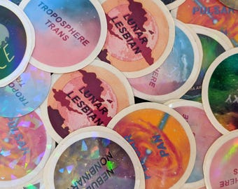Peut inclure: Une collection d'autocollants colorés avec différents motifs et textes sur le thème de l'espace, notamment "Space Ace", "Lunar Lesbian", "Troposphere Trans", "Galaxy Gay" et "Nebula Nonbinary".