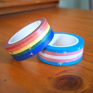 Puede incluir: Dos rollos de cinta adhesiva decorativa. Uno presenta un diseño de arcoíris con detalles de estrellas. El otro tiene un patrón de rayas azules, rosas y blancas con detalles de estrellas. Ambos rollos están sobre una superficie de madera.