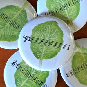 Puede incluir: Cuatro botones redondos blancos con un diseño de hoja verde y notas musicales impresas en ellos. Las hojas son de un verde claro con un patrón de venas verde oscuro. Las notas musicales son negras.