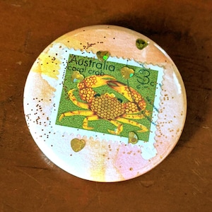 May include: A round, white button with a pink and orange watercolor background. A vintage Australian postage stamp with a coral crab is adhered to the button. The stamp says "Australia coral crab 3c".