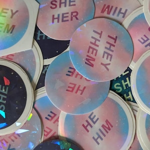 Pode incluir: Uma coleção de adesivos iridescentes e coloridos com várias combinações de pronomes, incluindo "they/them", "he/him", "she/her" e "he/she". Os adesivos têm um design de galáxia com cores rosa, azul e branco.