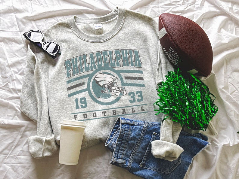 May include: A grey sweatshirt with a vintage-style graphic of the Philadelphia Eagles logo, the team name, and the numbers 19 and 33. The sweatshirt is laid out on a white surface with a football, a green pom-pom, a pair of sunglasses, and a white takeaway cup.