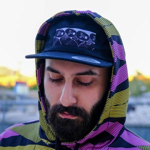 May include: A person wearing a black and yellow striped hooded sweatshirt with a black baseball cap. The cap has a white embroidered design of four faces.