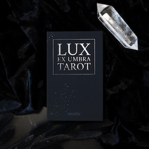 May include: A black tarot card deck with the words "LUX EX UMBRA TAROT" in silver lettering. A clear crystal point is positioned above the deck. The word "mystiic" is printed at the bottom of the deck.