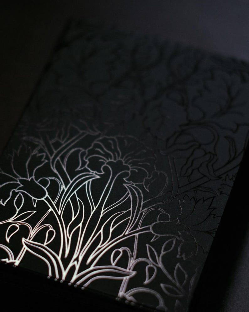 May include: A black notebook with a floral design. The design is a white, stylized illustration of flowers and leaves. The notebook is rectangular and appears to be made of a textured material.