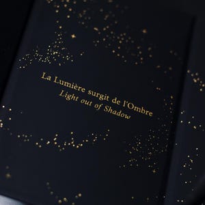 May include: Dark blue book with gold foil accents. The cover features scattered gold dots and the text "La Lumi&egrave;re surgit de l'Ombre Light out of Shadow" in gold lettering. The book's design evokes a starry night sky.
