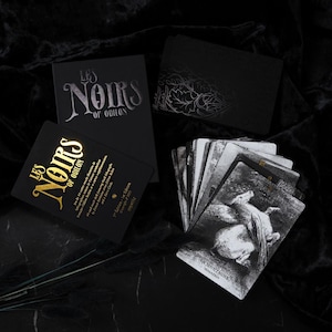 以下が含まれることがあります： タロットカードと箱のセット。箱は黒色で、「LES NOIRS OF ODILON」と銀色と金色の文字で書かれています。カードは白黒のイラストが特徴です。全体的な美学は暗く神秘的です。