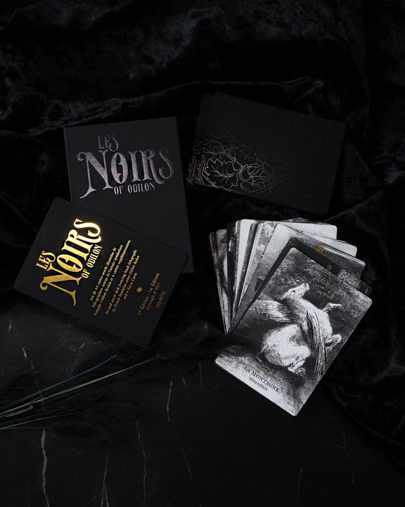 May include: A collection of tarot cards and boxes. The boxes are black with silver and gold lettering that reads "LES NOIRS OF ODILON." The cards feature black and white illustrations. The overall aesthetic is dark and mysterious.