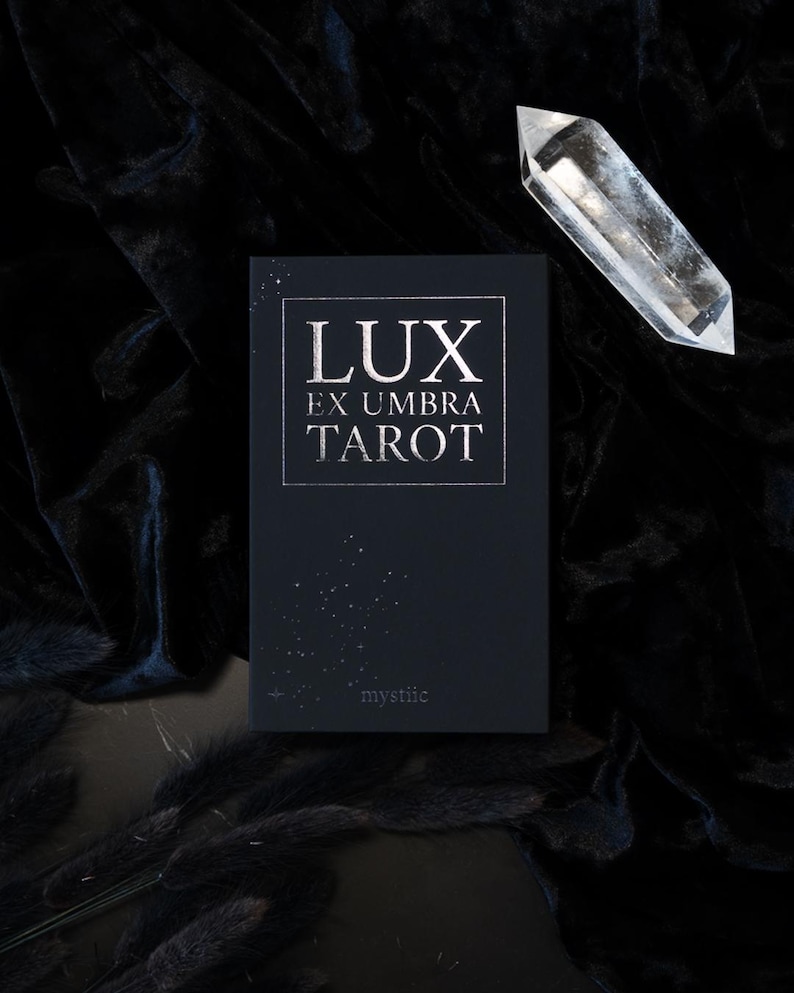 May include: A black tarot card deck with the words "LUX EX UMBRA TAROT" in silver lettering. A clear crystal point is positioned above the deck. The word "mystiic" is printed at the bottom of the deck.