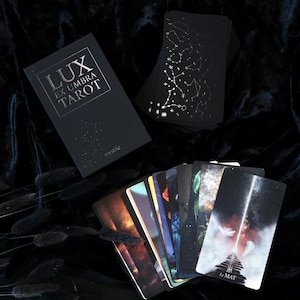 Op de afbeelding: Een zwart tarotdeck met gouden sterrenbeeldontwerpen en de tekst "LUX EX UMBRA TAROT" op de doos. Verschillende kaarten zijn uitgespreid en tonen kleurrijke illustraties. Het deck is gelabeld "mystic".
