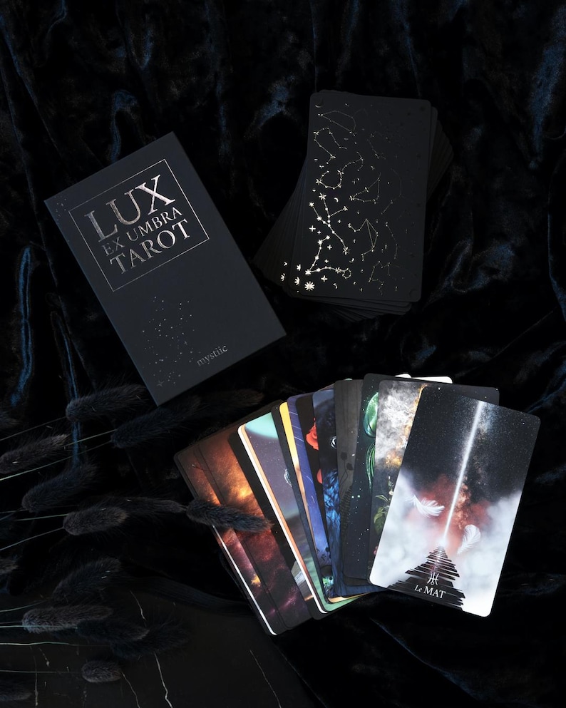 May include: A black tarot deck with gold constellation designs and the text "LUX EX UMBRA TAROT" on the box. Several cards are fanned out, displaying colorful illustrations. The deck is labeled "mystic".