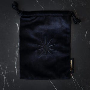 May include: A dark navy blue velvet drawstring bag with an embroidered starburst design. The bag has a small tag with the word "mystic" in gold lettering. The bag is closed with a black cord.
