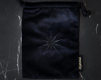 May include: A dark navy blue velvet drawstring bag with an embroidered starburst design. The bag has a small tag with the word "mystic" in gold lettering. The bag is closed with a black cord.