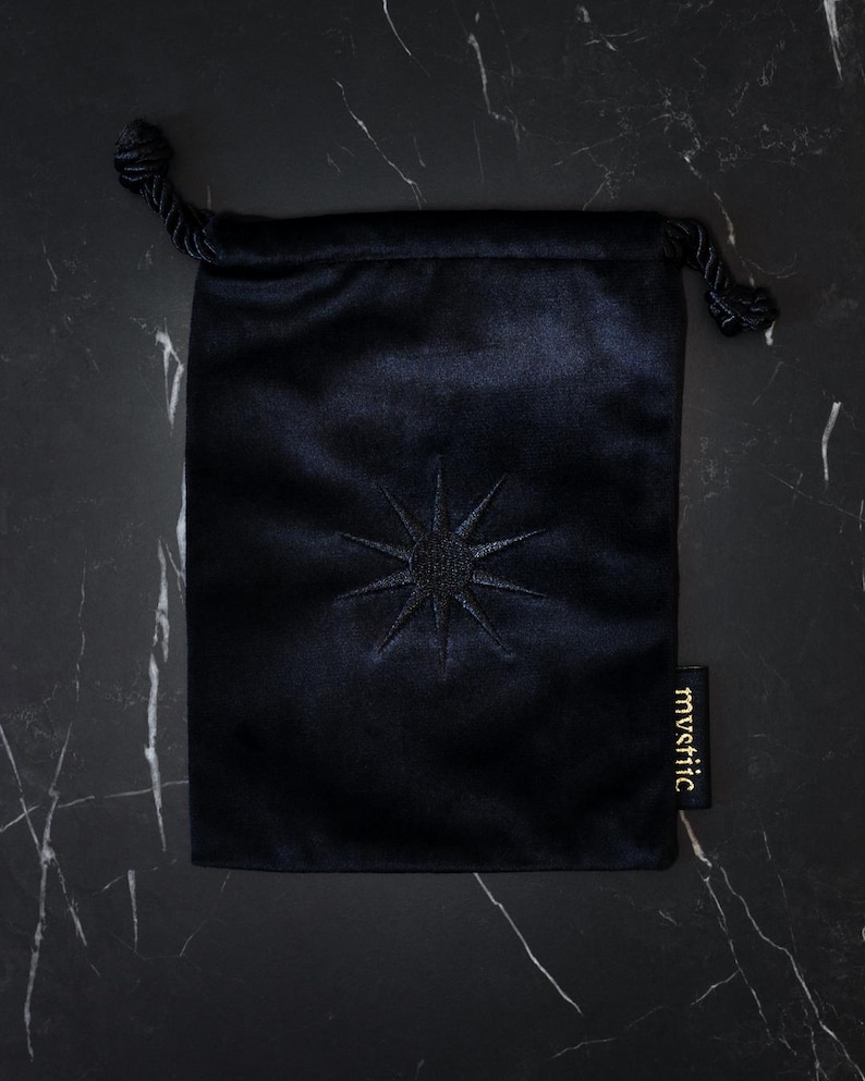 May include: A dark navy blue velvet drawstring bag with an embroidered starburst design. The bag has a small tag with the word "mystic" in gold lettering. The bag is closed with a black cord.