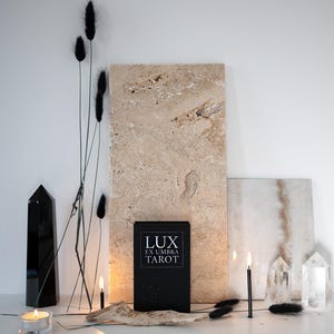 May include: A still-life arrangement featuring a black tarot card box with the text "LUX EX UMBRA TAROT", a black crystal, candles, dried black flowers, and clear quartz crystals. The background includes beige stone slabs.