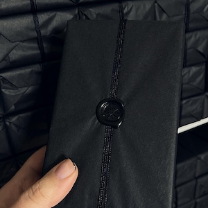 May include: A black rectangular package wrapped in black paper, secured with a black cord and sealed with a black wax stamp. The package is held by a person. Other wrapped packages are in the background.