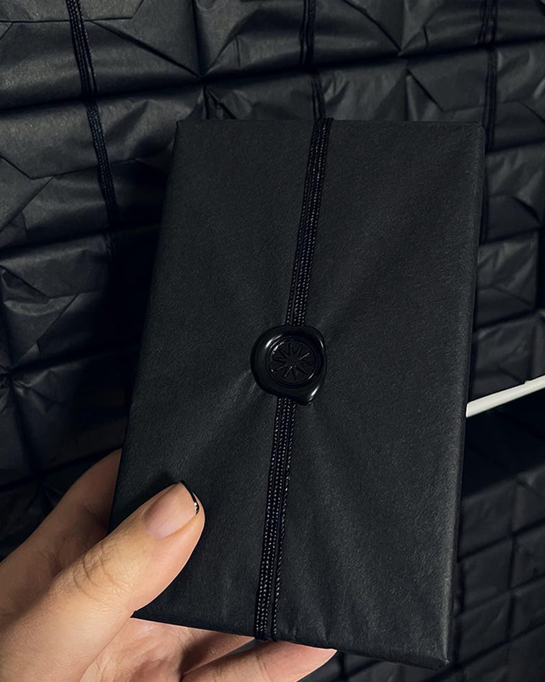 May include: A black rectangular package wrapped in black paper, secured with a black cord and sealed with a black wax stamp. The package is held by a person. Other wrapped packages are in the background.