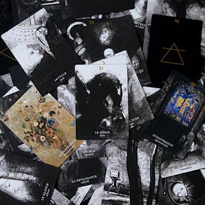 May include: A collection of tarot cards with black and white illustrations and French text. The cards feature images of figures, symbols, and text, with a few cards displaying gold and blue accents. The cards are scattered across a dark surface.