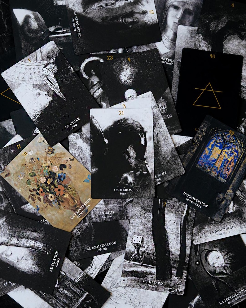 May include: A collection of tarot cards with black and white illustrations and French text. The cards feature images of figures, symbols, and text, with a few cards displaying gold and blue accents. The cards are scattered across a dark surface.