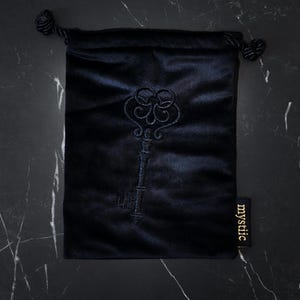 May include: A black velvet drawstring bag with an embroidered key design. The bag has a tag that reads "mystiic". The key design is dark blue and the bag is on a black marble surface.