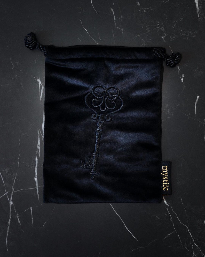 May include: A black velvet drawstring bag with an embroidered key design. The bag has a tag that reads "mystiic". The key design is dark blue and the bag is on a black marble surface.
