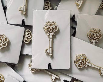 May include: Gold-toned key-shaped pins with white enamel detailing are displayed on white cards. The pins have an ornate design and are packaged in clear plastic. The background is filled with similar pins, suggesting a collection or bulk sale.