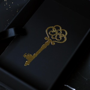 May include: A gold-colored, ornate key design is printed on a black surface, possibly a box or card. The key has a detailed, decorative head and a long stem. The background is dark, with subtle highlights.