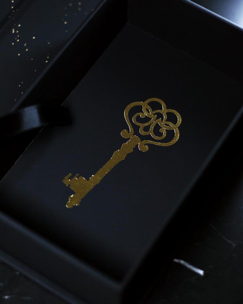 May include: A gold-colored, ornate key design is printed on a black surface, possibly a box or card. The key has a detailed, decorative head and a long stem. The background is dark, with subtle highlights.