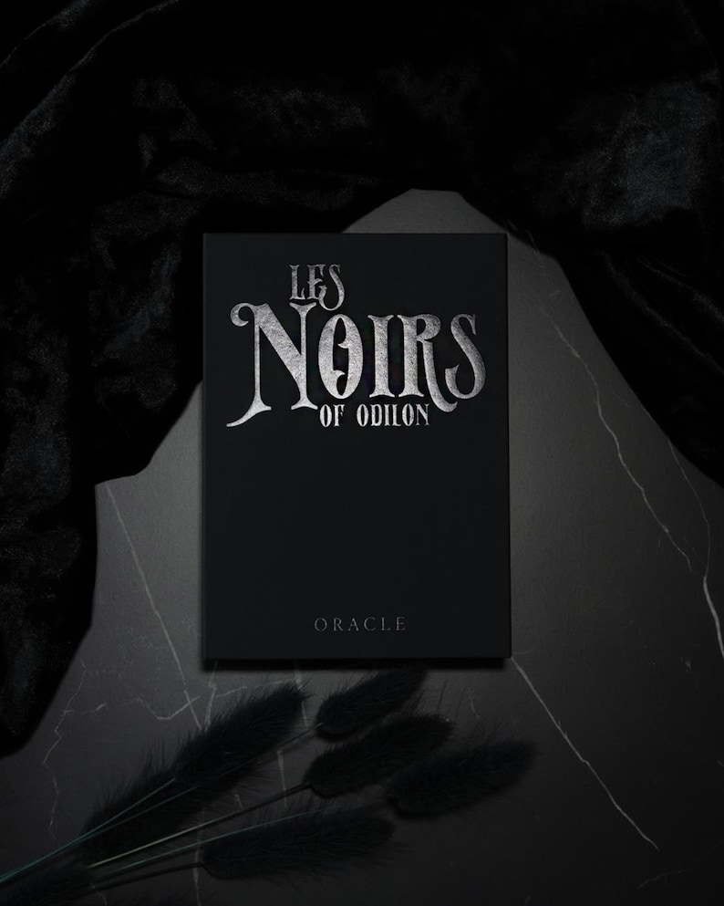 May include: A black box with silver lettering that reads "LES NOIRS OF ODILON" and the word "ORACLE" below. The box is on a dark surface with black accents.